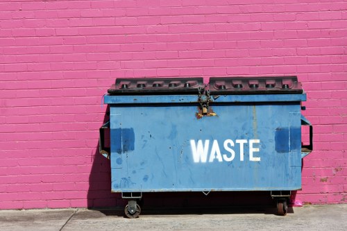 A large blue waste bin with a hinged lid, positioned on a concrete pavement adjacent to a vibrant pink brick wall. The bin is made of metal or heavy-duty plastic with visible scratches and dirt marks, and the word 'WASTE' is printed in white uppercase letters on its front. The lid appears slightly weathered with some rust and grime around the locking mechanism. The bin has four small wheels at its base, allowing for mobility, and is situated in a clean, outdoor urban environment, possibly in a residential or commercial area within Seven Sisters or nearby London postcodes. The scene is lit by natural daylight, casting a distinct shadow to the left of the bin, emphasizing its three-dimensional form. This image is representative of rubbish collection and disposal services provided by Commercial Waste Seven Sisters, highlighting waste management infrastructure common to local businesses and properties in the area.