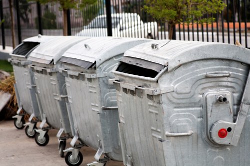A row of four large, open-top metal wheelie bins made of galvanized steel is positioned on a paved surface, with each bin featuring black plastic lids tilted back to reveal their empty interiors. The bins are lined up close together, displaying a uniform design with visible ridges and a textured finish. Each bin is equipped with four caster wheels at the base for mobility, and the front of the bins includes a lockable latch mechanism to secure the lids. A small round red reflector or warning light is attached to the right side of the nearest bin. In the background, there is a black metal fence, some trees with green foliage, and a glimpse of parked vehicles, suggesting an urban or suburban setting possibly near commercial premises or a waste collection point around Seven Sisters. The lighting is natural, indicating the image was taken during daylight hours, and the overall scene emphasizes waste storage and collection infrastructure suitable for professional rubbish removal services in the local area.