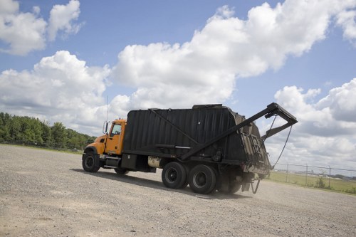A large industrial waste collection truck belonging to Commercial Waste Seven Sisters is parked on a vast gravel surface under a partly cloudy sky. The vehicle is painted in bright orange and has a black body designed for rubbish collection, with a hydraulic lift system at the rear which is currently in an elevated position, indicating active loading or unloading of waste materials. The truck's wheels are sturdy and black, suited for heavy-duty use, and the dirty gravel ground suggests a temporary or construction site environment. In the background, there are distant fields and some trees, hinting at a location near Seven Sisters in North London or Essex. The overall scene emphasizes the operational aspect of commercial rubbish removal services, with the truck ready for waste collection or disposal, consistent with the services offered by Commercial Waste Seven Sisters for local businesses and facilities in the area.