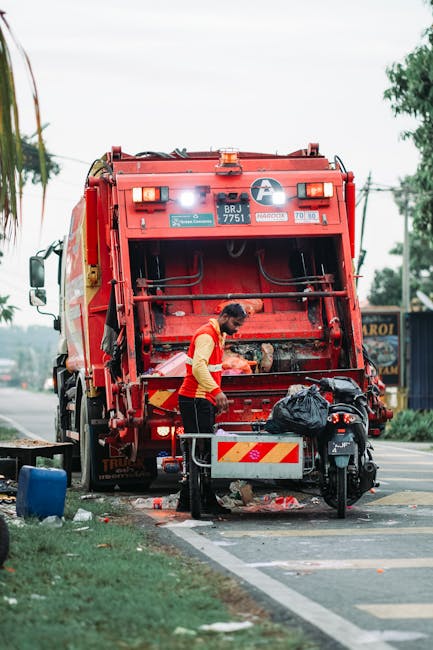 A street scene showing a red rubbish collection truck parked near the curb, with its rear compartment open, revealing internal mechanisms for waste disposal. A worker wearing a high-visibility vest an