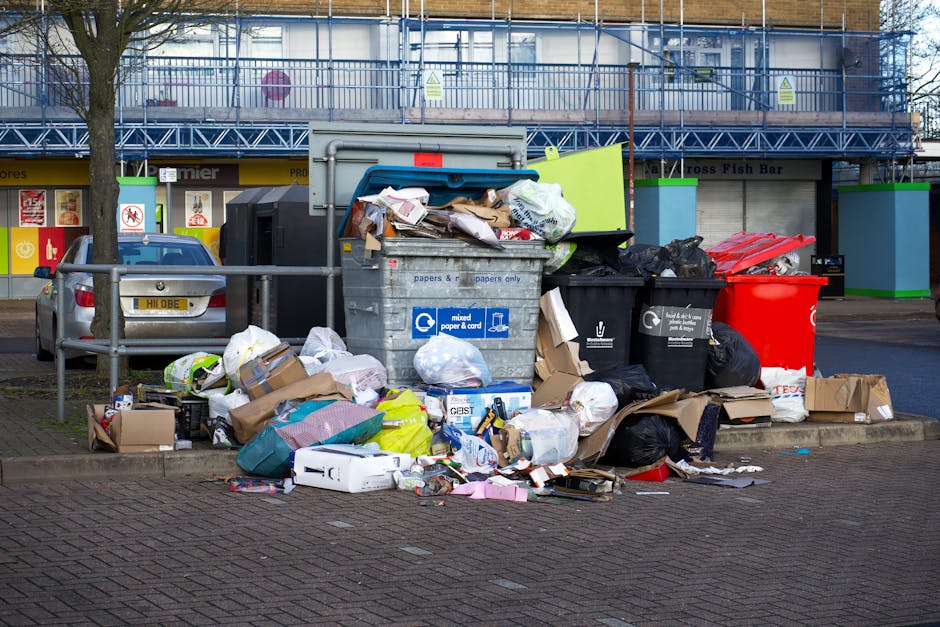 A collection of overflowing rubbish bins and scattered waste materials on a paved urban parking area. The scene features various black, grey, and red bin containers, some with their lids open, reveali