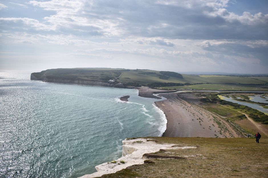 A wide view of a coastal landscape showing a sandy beach with several wooden groynes extending into the calm, light turquoise sea under a partly cloudy sky. In the foreground, a series of pathways and